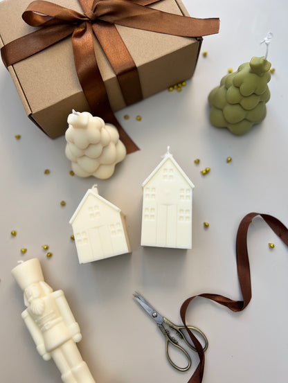 Decorative candles shaped like houses, nutcracker and christmas trees, a gift box with a brown ribbon, and scissors on a light surface.