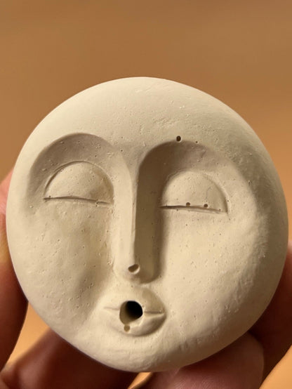 Hand holding a moon face shaped incense holder with a central hole against a blurred background